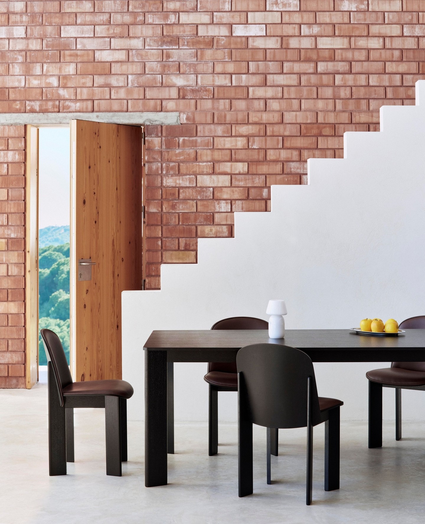 Modern dining room with black table and chairs, stepped white plaster and exposed red brick, open wooden door to green view