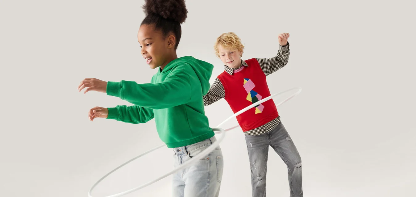 Una ragazza con una felpa verde con cappuccio fa hula hoop mentre un ragazzo con un gilet rosso guarda e si equilibra su un piede su uno sfondo semplice.