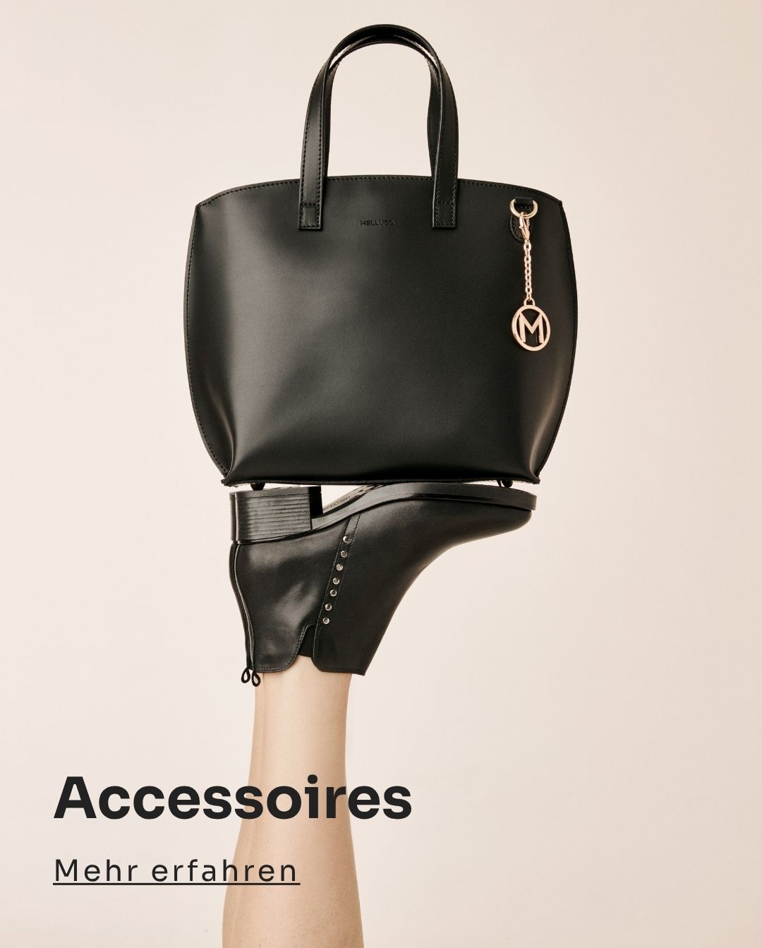 Black leather tote bag with gold 'M' charm balanced on a foot wearing a black ankle boot against a neutral background