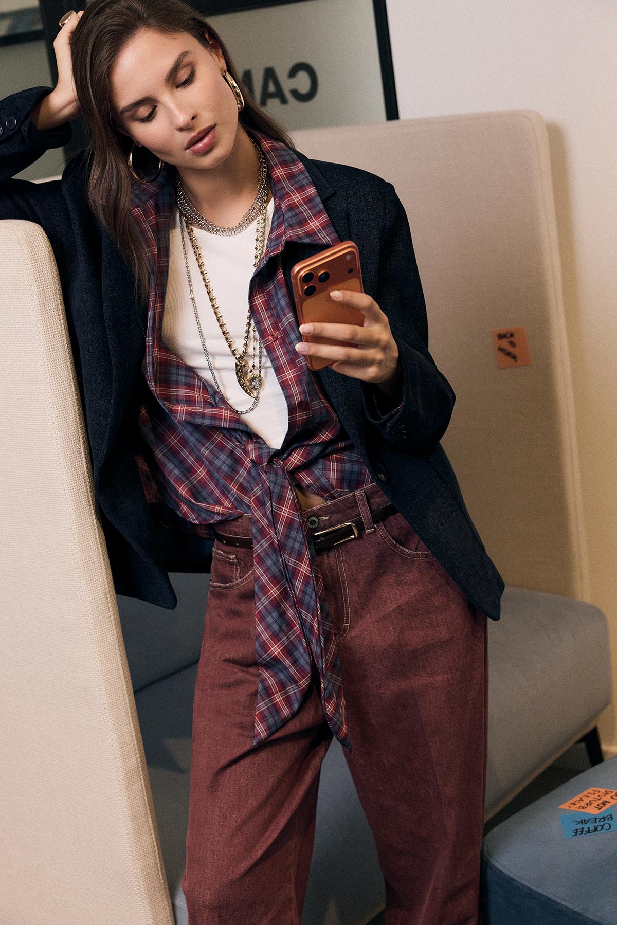 Young woman seated on sofa checking smartphone, wearing layered plaid shirt, white tee, navy blazer, burgundy trousers and layered chains