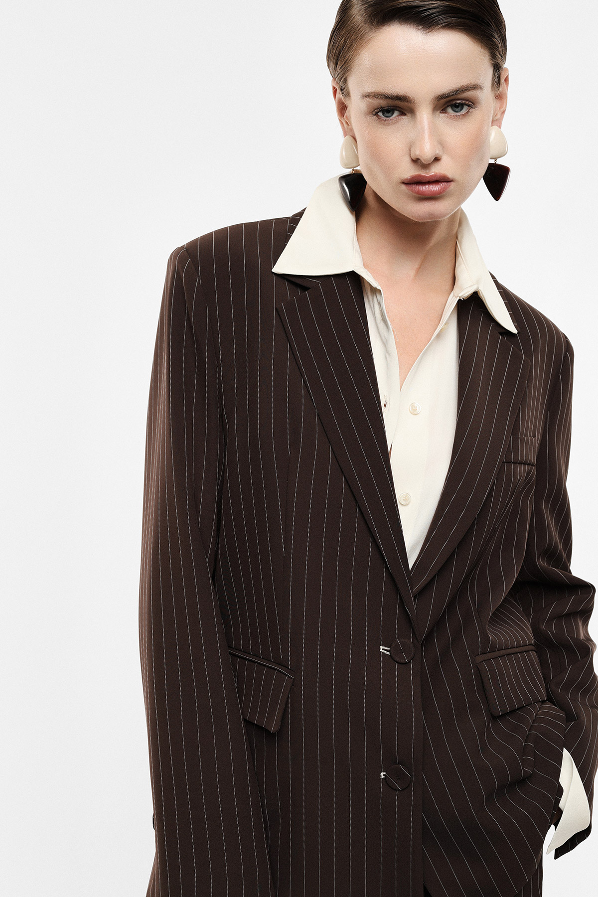 Model wearing a brown pinstripe blazer over a cream blouse with geometric statement earrings, hand in pocket, fashion portrait