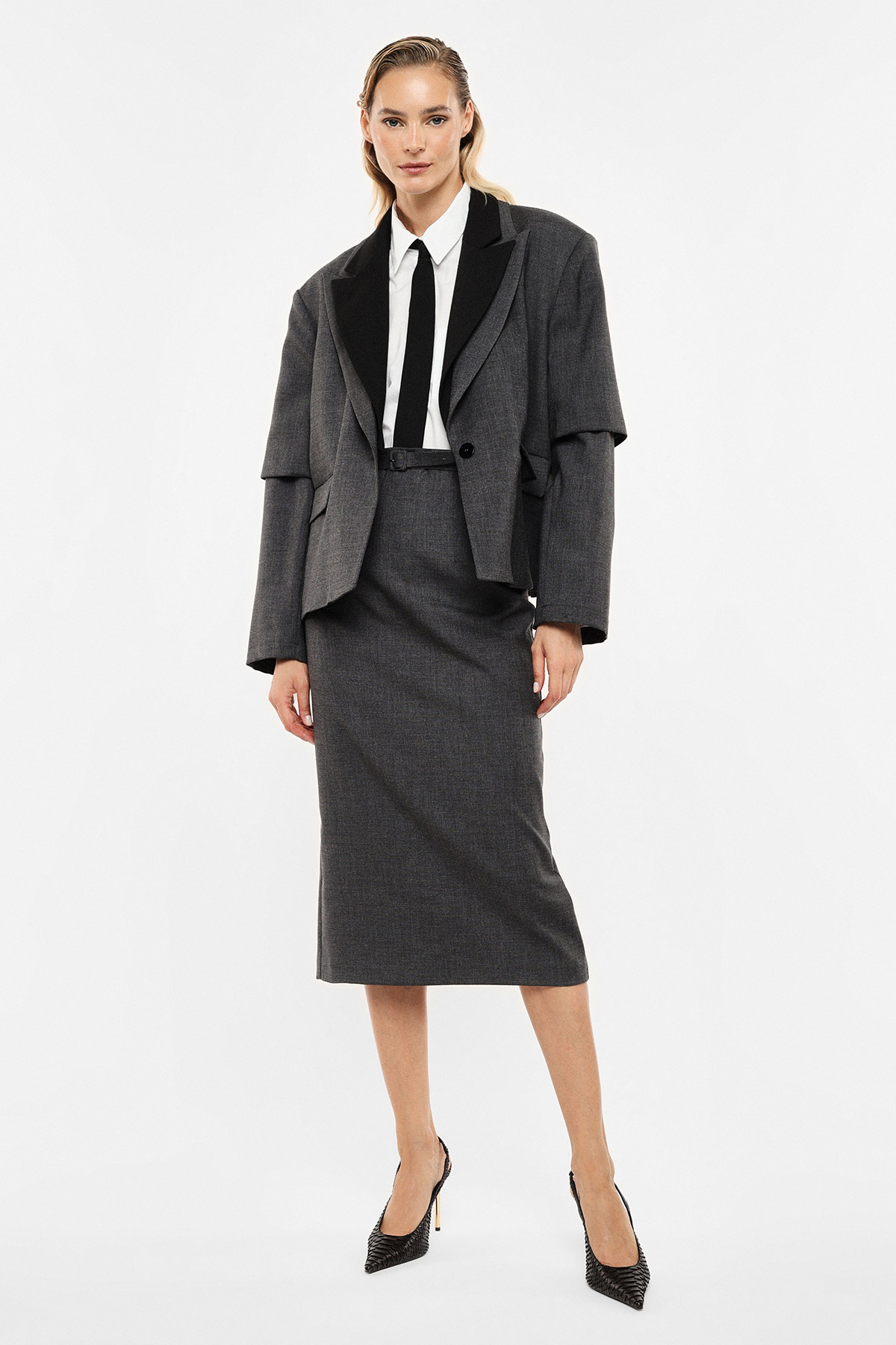 Woman modelling grey tailored skirt suit with black lapels, white shirt and narrow black tie, black heels on white background