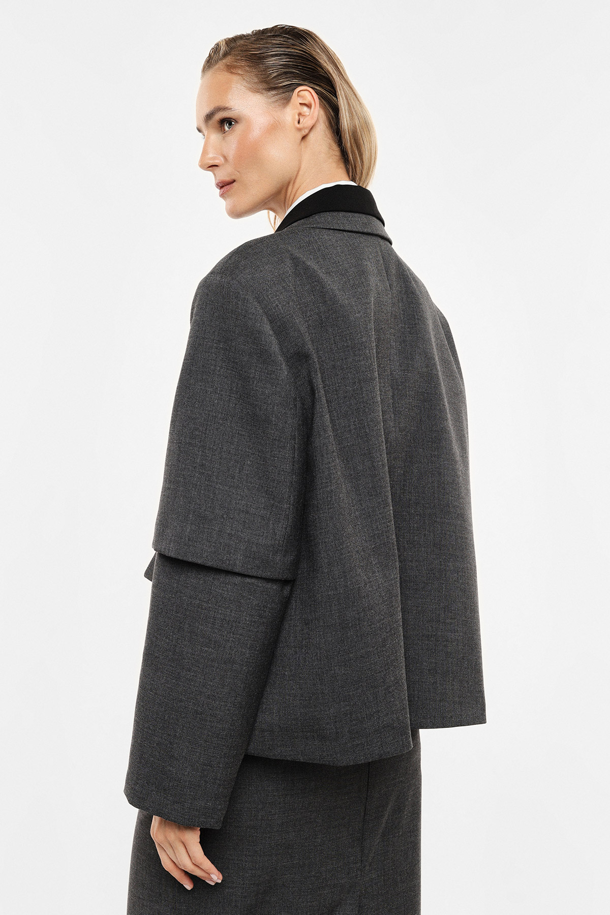 Woman in dark grey layered wool jacket viewed from back, looking over shoulder; tailored cropped coat for autumn/winter