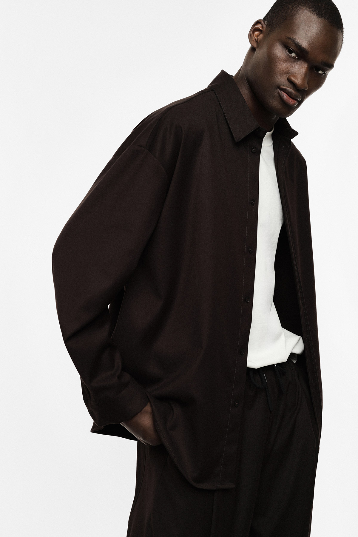 Model wearing oversized dark brown shirt over white T-shirt and loose trousers, hands in pockets, leaning forward against plain light background