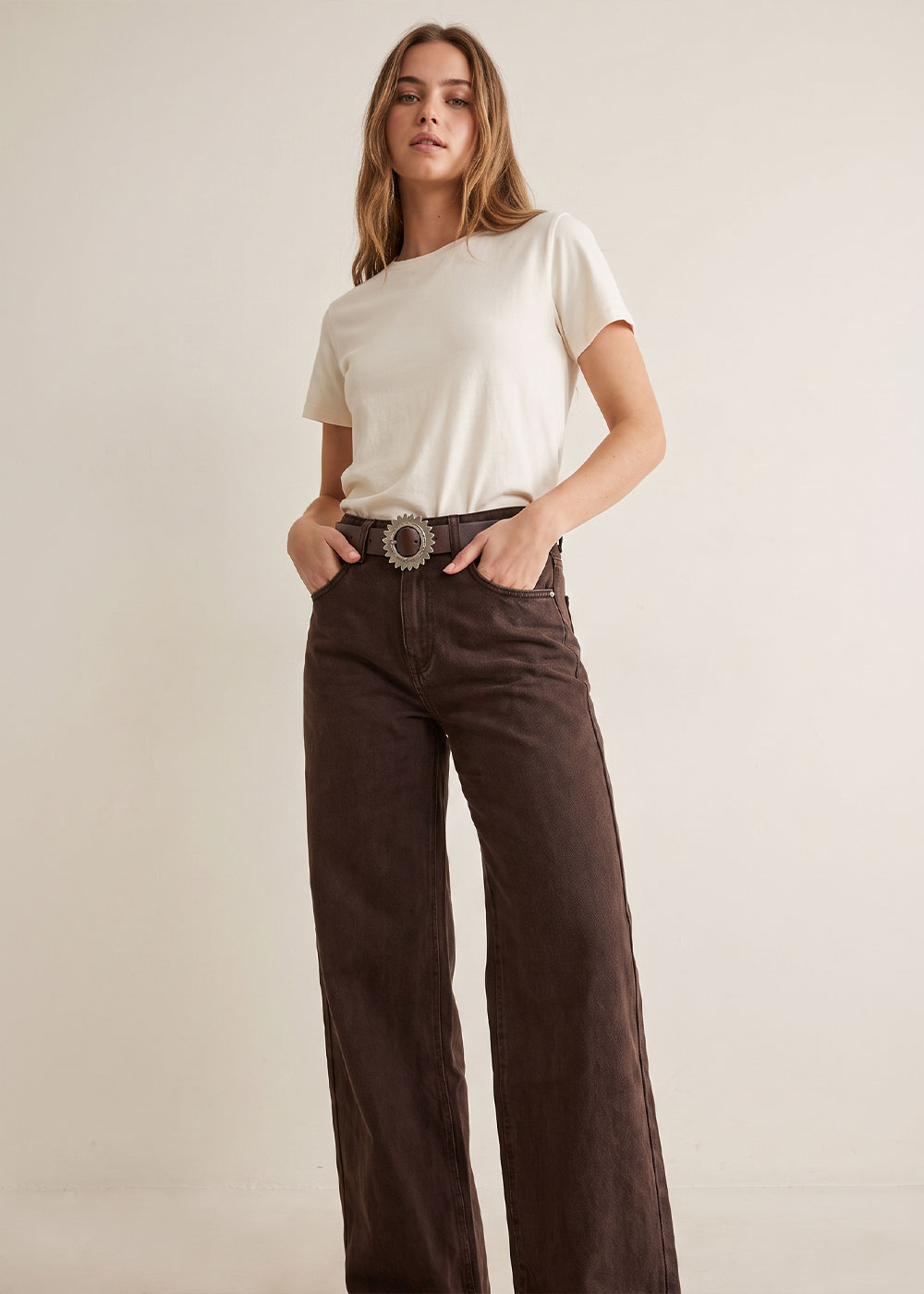 Model in cream short-sleeve t-shirt and high-waisted brown wide-leg jeans with statement belt, hands in pockets, standing against neutral background