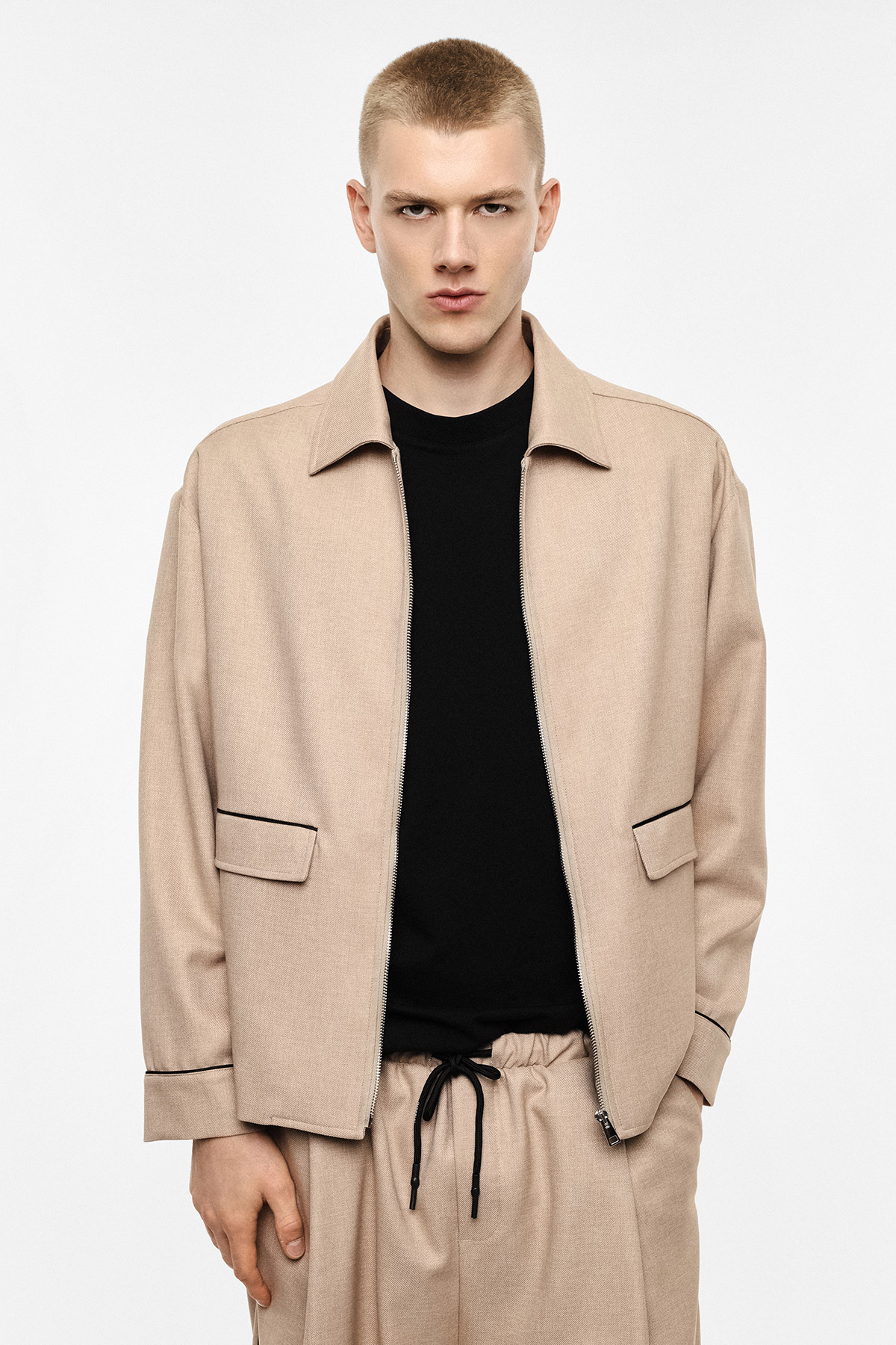 Young male model modelling a beige jacket and matching drawstring trousers with a black top, standing against a white background