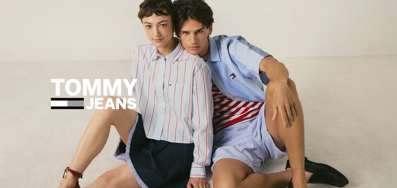 Due giovani persone sono sedute sul pavimento appoggiate l'una all'altra, indossando camicie a righe casual, pantaloncini e mocassini.