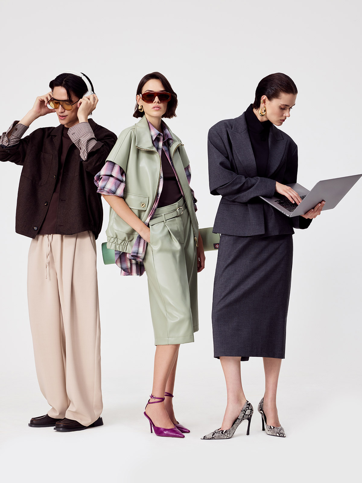 Three models: left with headphones and wide trousers; centre in green leather co‑ord and purple heels; right in grey skirt suit with laptop