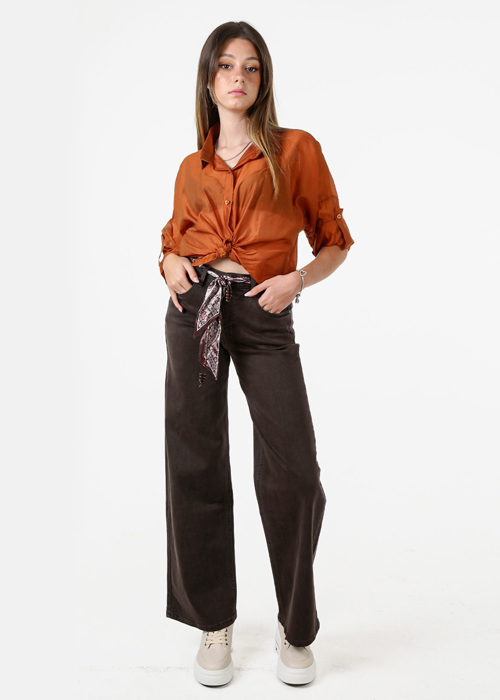 Full-length image of a woman modelling an autumn outfit — rust orange tied blouse, dark brown wide-leg trousers, patterned scarf belt and white platform trainers on a white background
