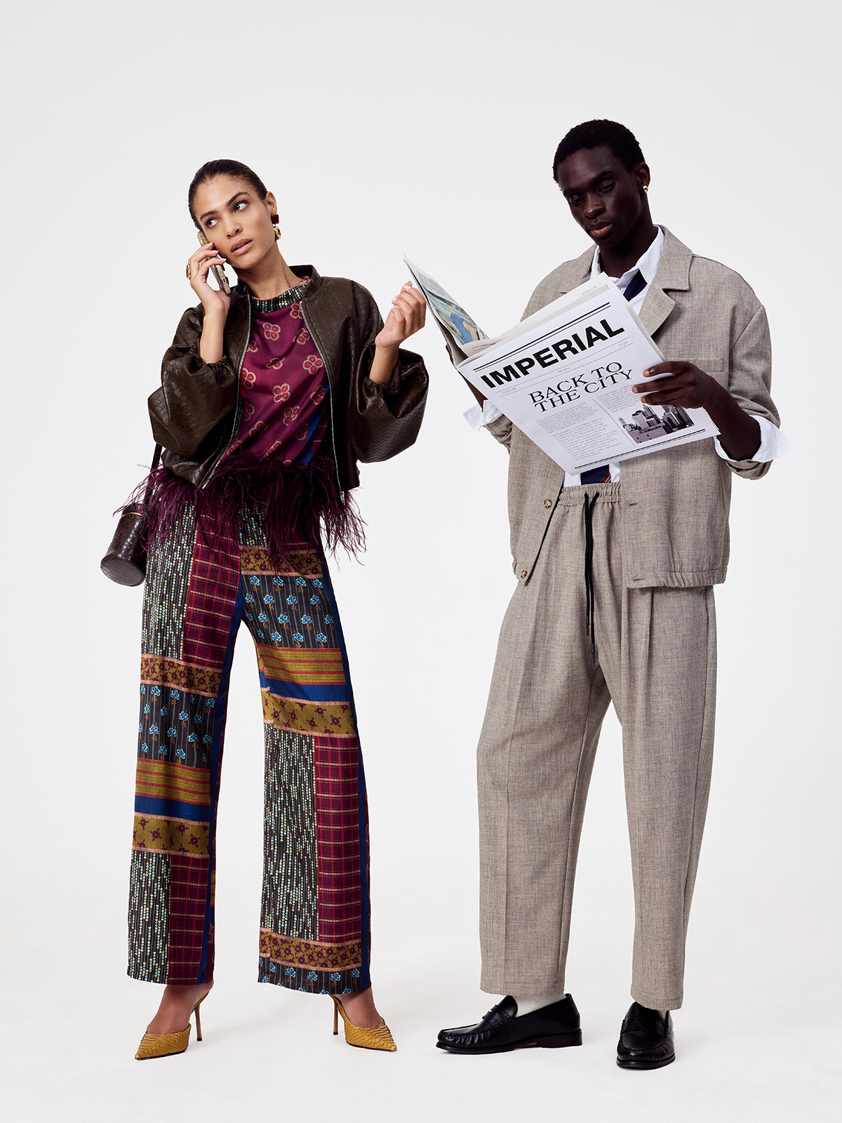 Two fashion models: woman in patterned patchwork trousers and feathered top on phone, man in relaxed beige suit reading a newspaper