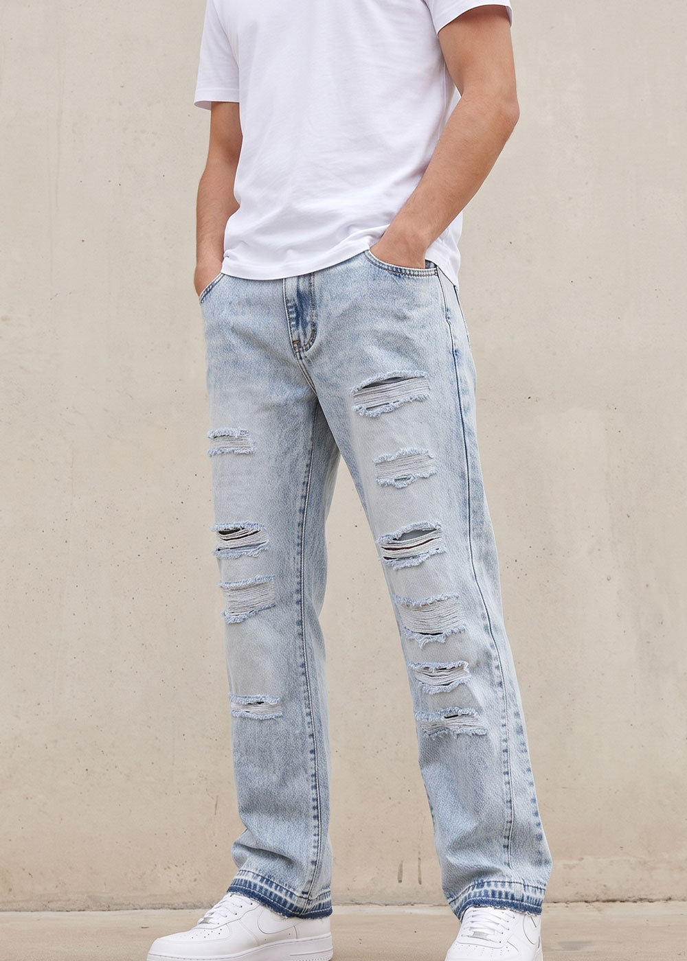 Person wearing a white T‑shirt, light blue distressed ripped straight‑leg jeans and white trainers, hands in pockets against a concrete wall
