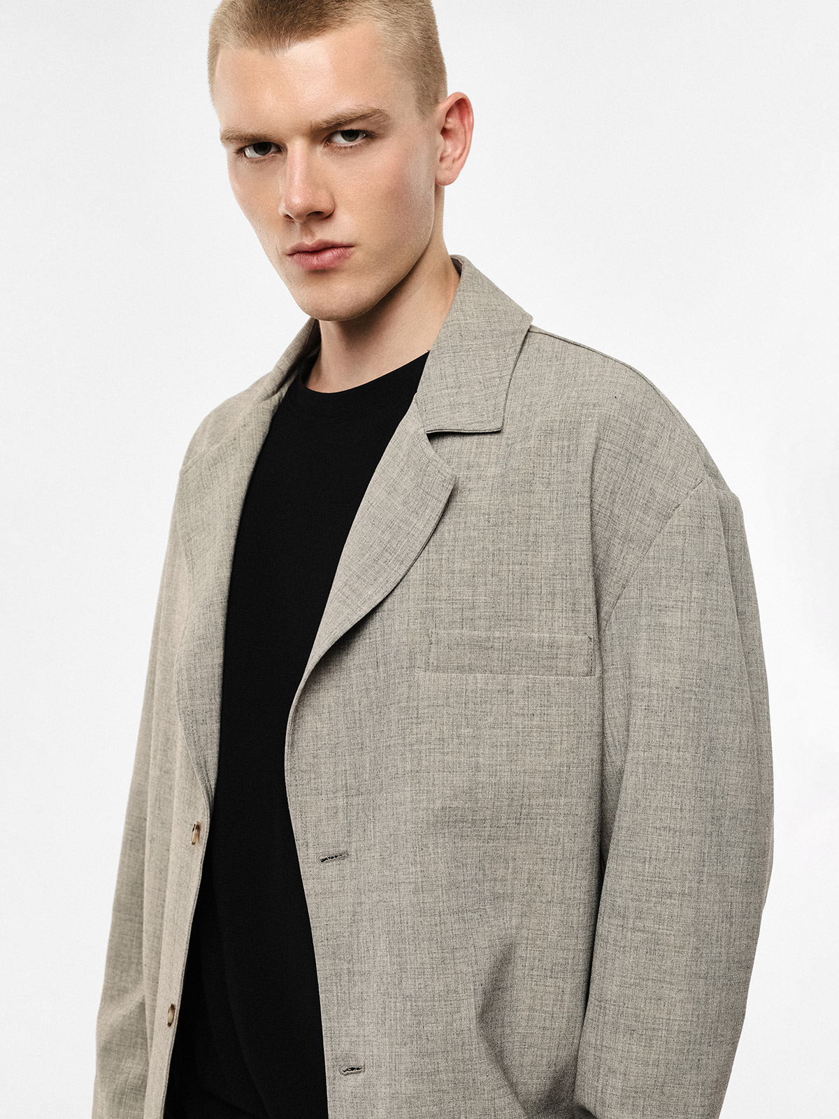 Male model wearing a light grey textured blazer over a black crew-neck top, three-quarter portrait on a plain white background with a serious expression