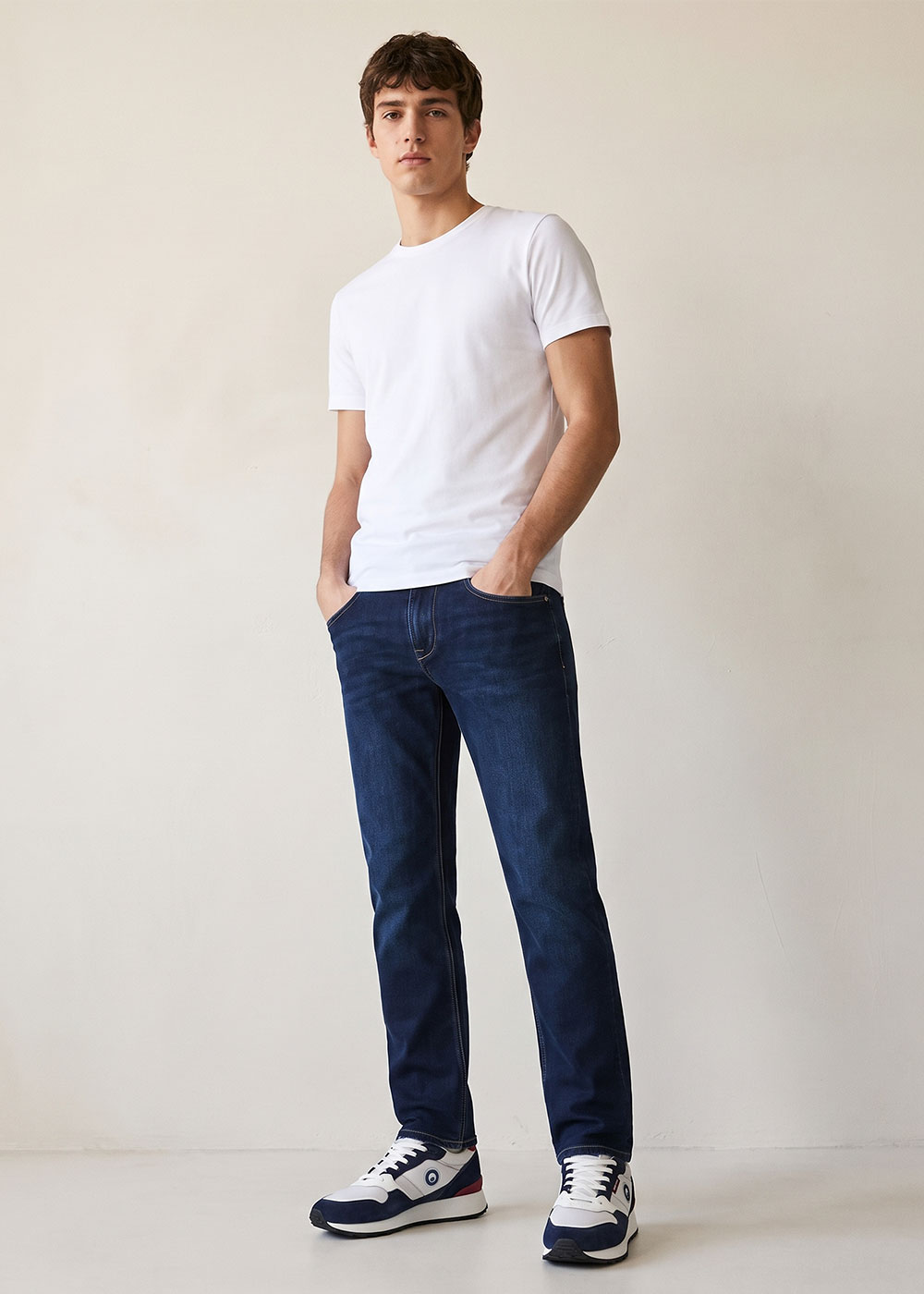 Full-length photo of a young man modelling a plain white T‑shirt, dark blue jeans and navy-white trainers against a neutral background