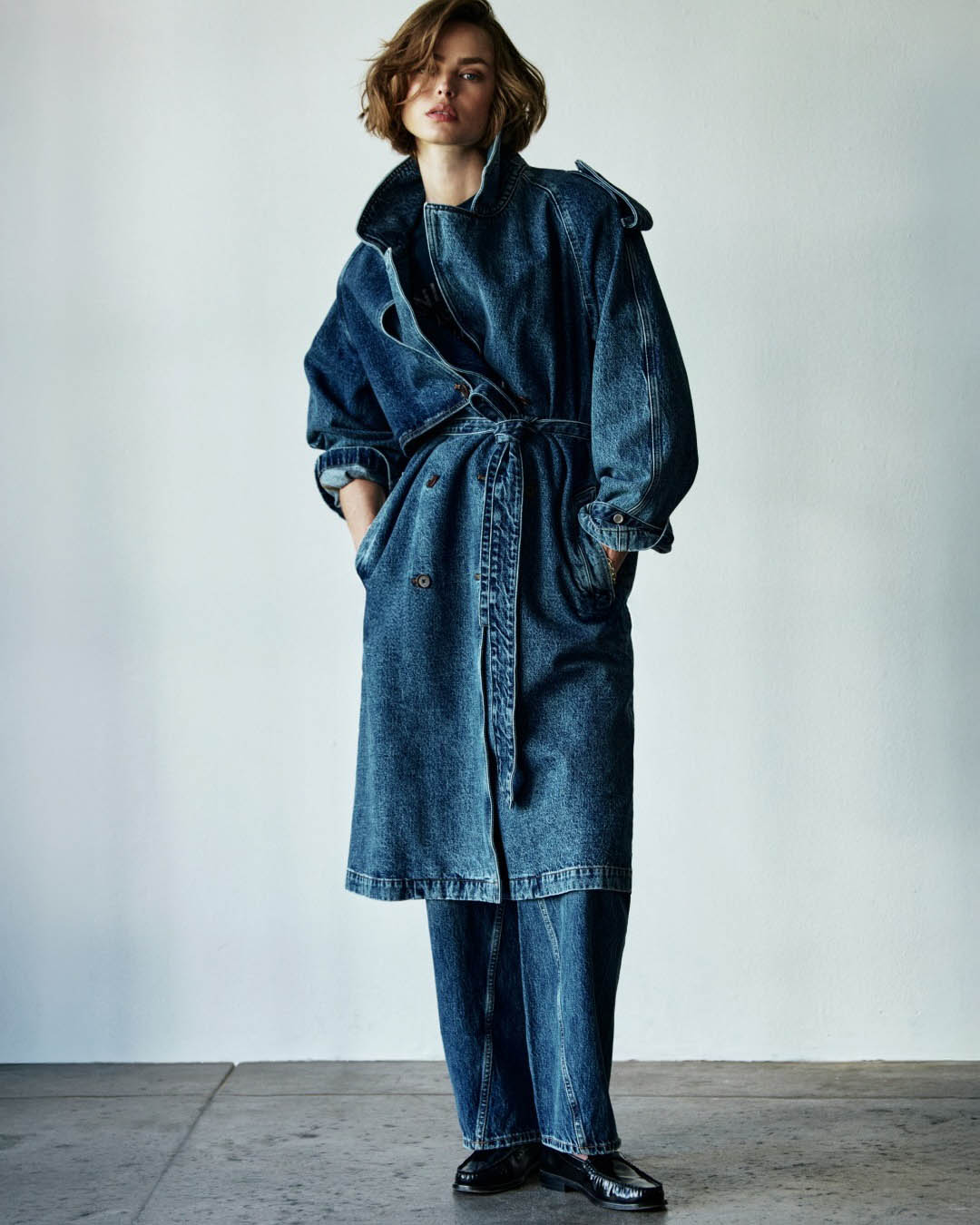 Model wearing oversized belted denim trench coat with matching wide-leg jeans and black loafers, standing against a plain light wall