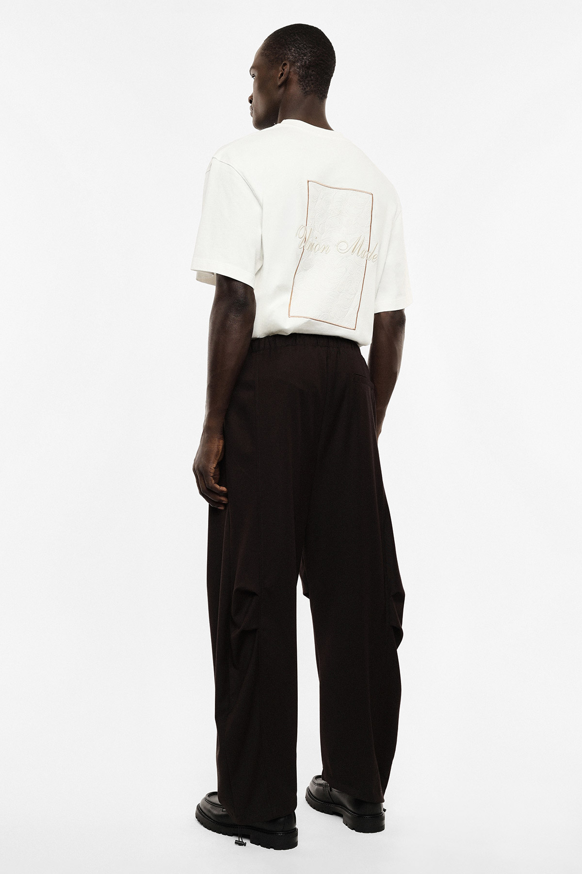 Rear view of male model wearing white oversized T‑shirt with stitched back patch, wide black trousers and black shoes