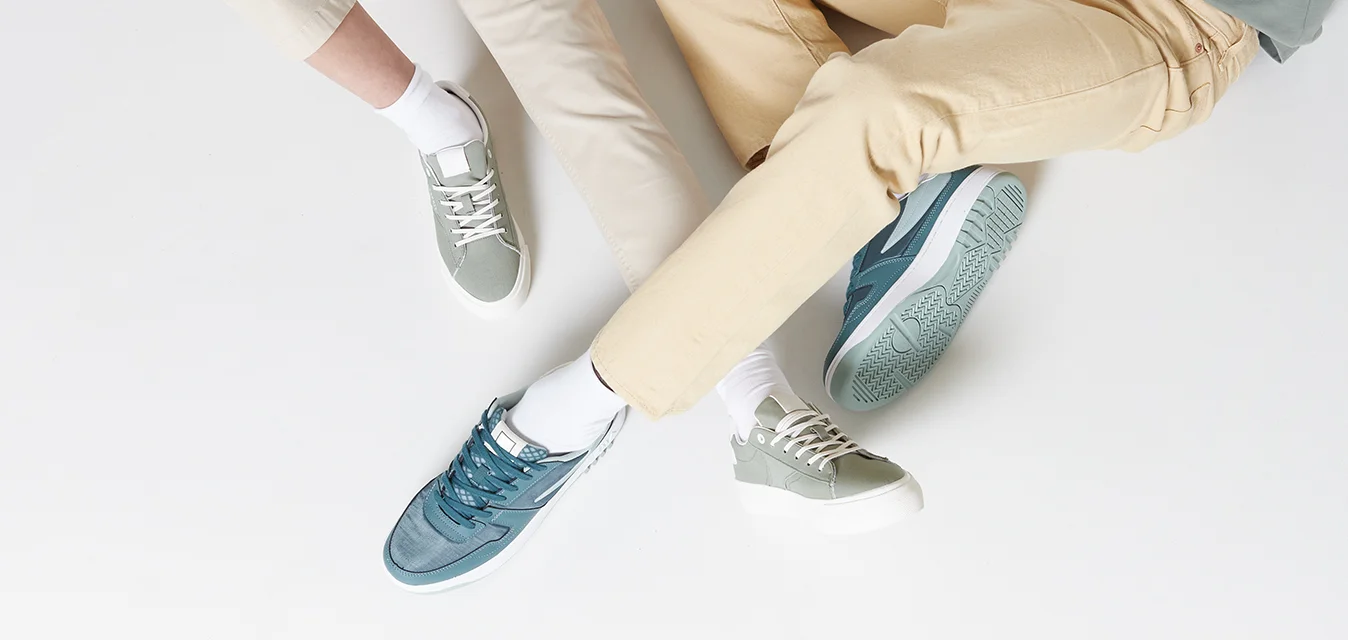 Due persone siedono su una superficie bianca, indossando una maglietta verde chiaro e pantaloni beige. Le calzature includono sneakers testurizzate bicolore in verde acqua e grigio.
