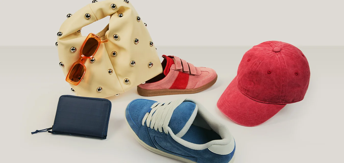 Borsa beige con borchie, occhiali da sole arancioni, sneakers rosa e blu, cappellino rosso e portafoglio nero su una superficie chiara.
