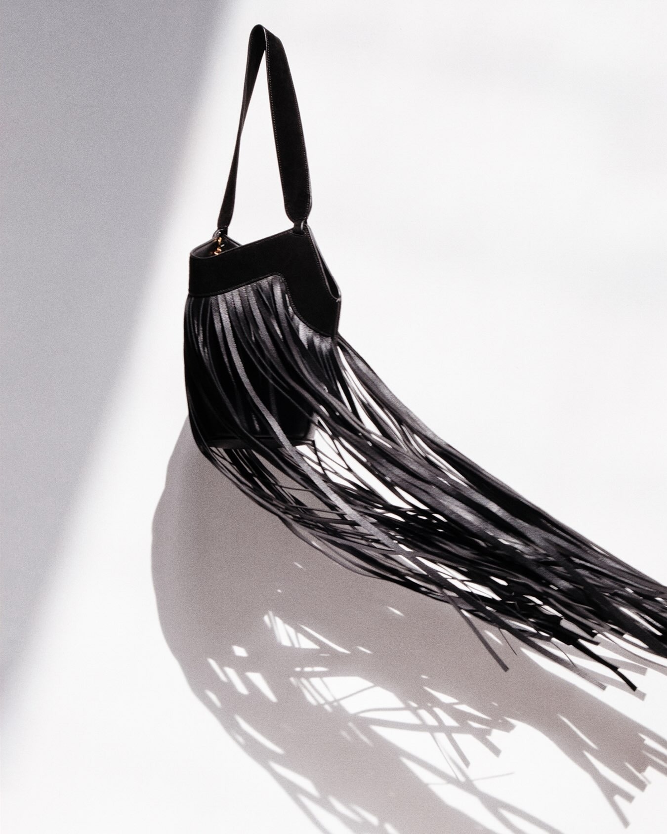 Black leather shoulder bag with long fringe detail hanging against a white background, dramatic shadow for fashion product imagery
