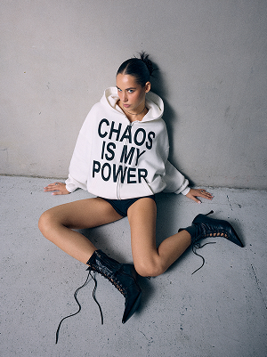 Donna con felpa bianca con cappuccio e scritta „CHAOS IS MY POWER“, shorts neri e stivali neri alti.