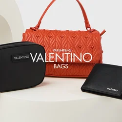 Tre accessori: una borsa arancione strutturata con manico superiore, un piccolo astuccio nero con cerniera e un'etichetta del marchio, e un portafoglio nero, tutti su un piedistallo.