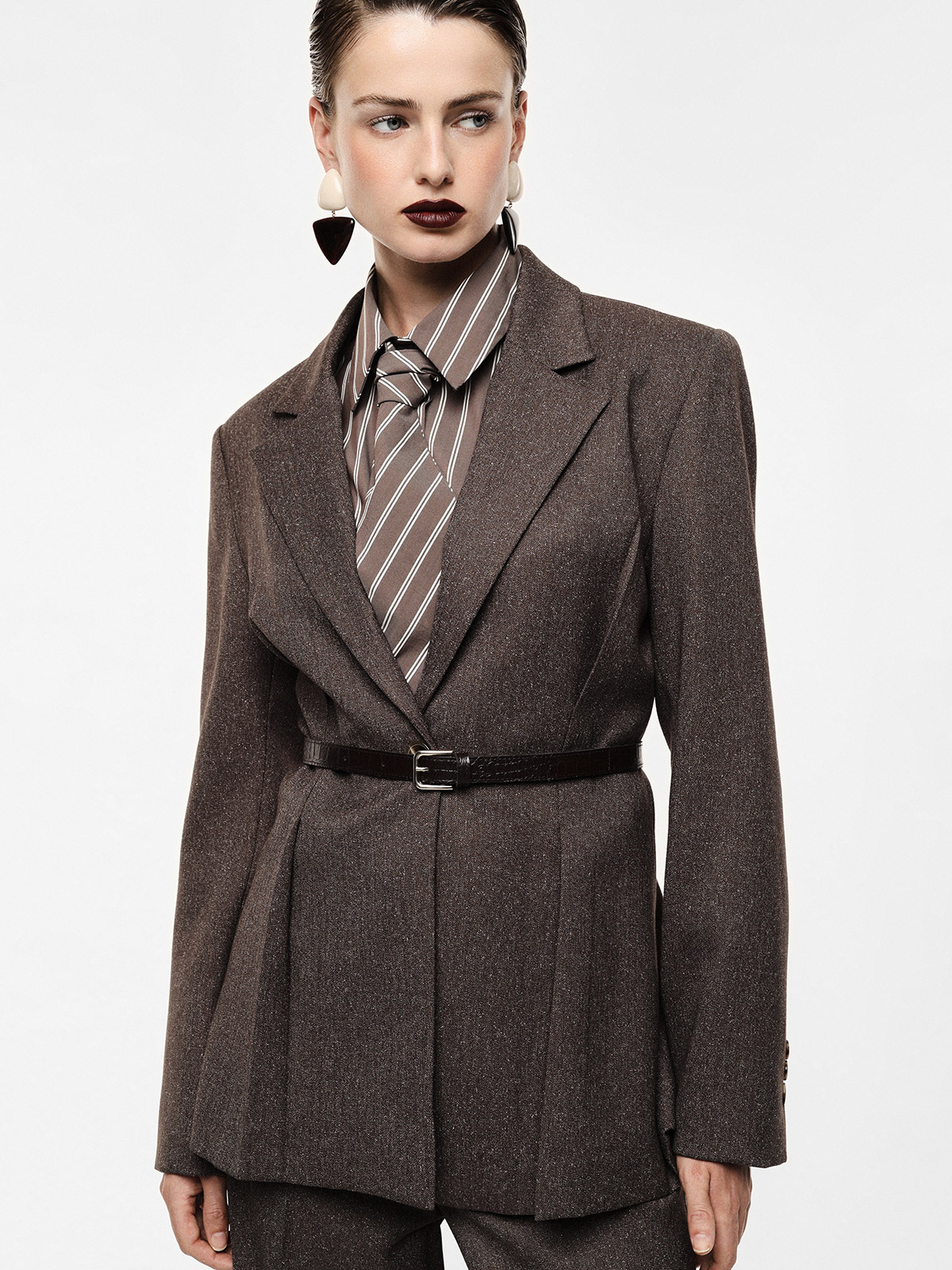 Female model wearing a brown tailored blazer cinched with a slim belt, striped tie and statement earrings, looking right on a white background