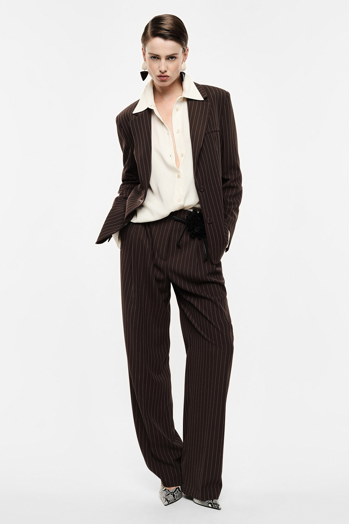 Woman wearing brown pinstripe suit with wide-leg trousers and cream blouse, hands in pockets on white background