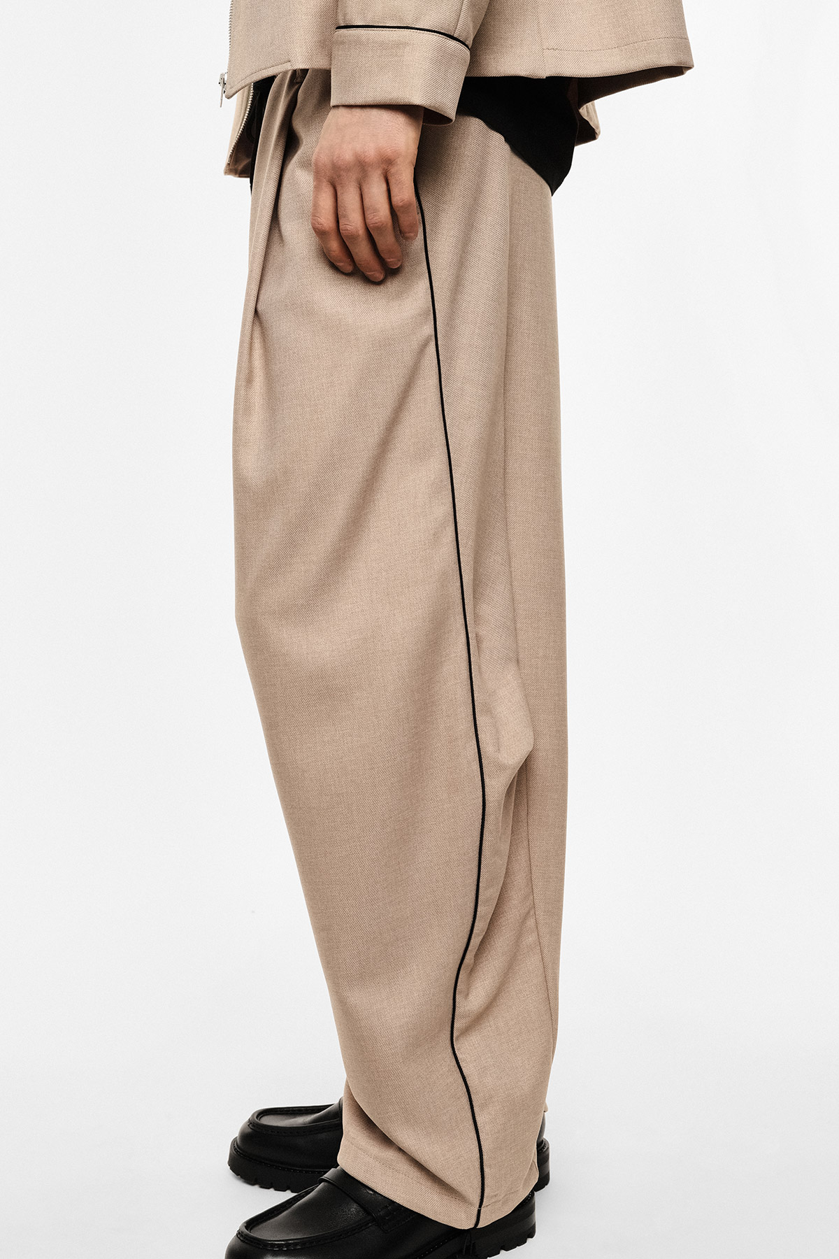 Model wearing beige wide-leg trousers with black side piping, hand by hip and black chunky leather shoes