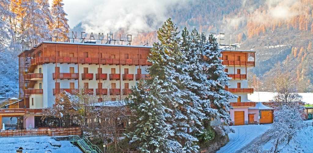 Snow-covered mountain hotel with wooden balconies beside tall snow-dusted pine trees and misty forested slopes