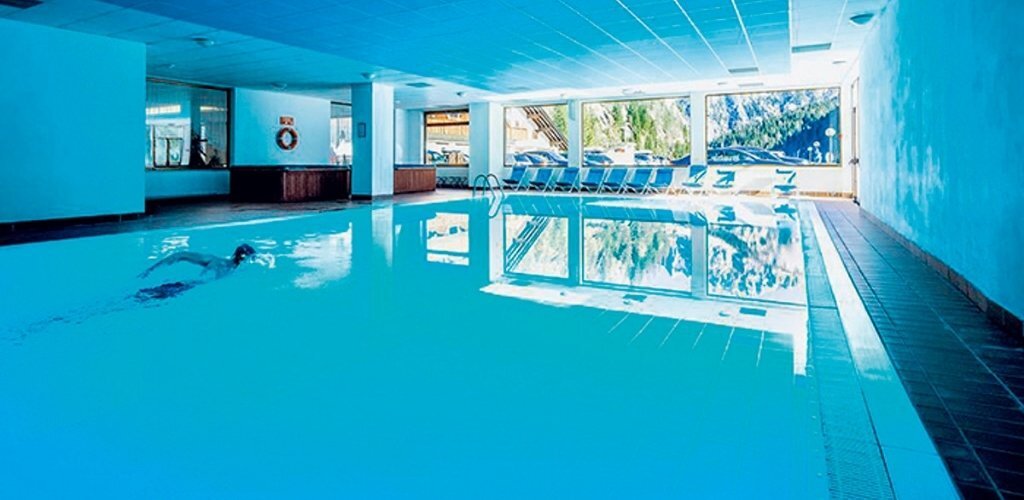 Indoor hotel swimming pool with loungers beside large windows overlooking mountain scenery, person swimming laps