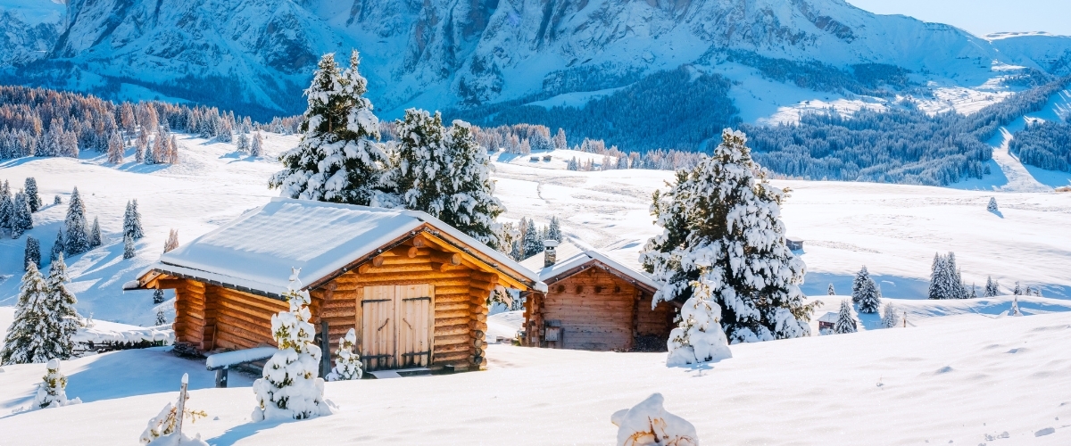 Snow-covered wooden cabins among pine trees on a sunlit alpine meadow with snow-capped mountains in the background