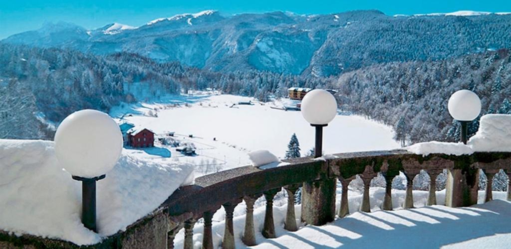 Snowy mountain lake view from a stone balcony with snow-covered globe lamps and a red cottage by the frozen water