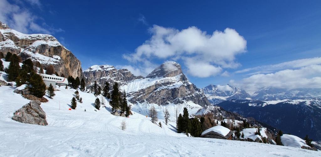 Snow-covered alpine slopes and pine trees with rocky mountain peaks under a blue sky, ski pistes and a small hut