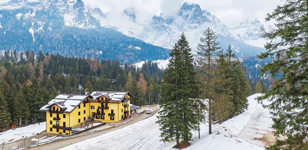 Yellow alpine chalet beside a snow-covered pine forest and winding road, with majestic snow-capped mountains in the background