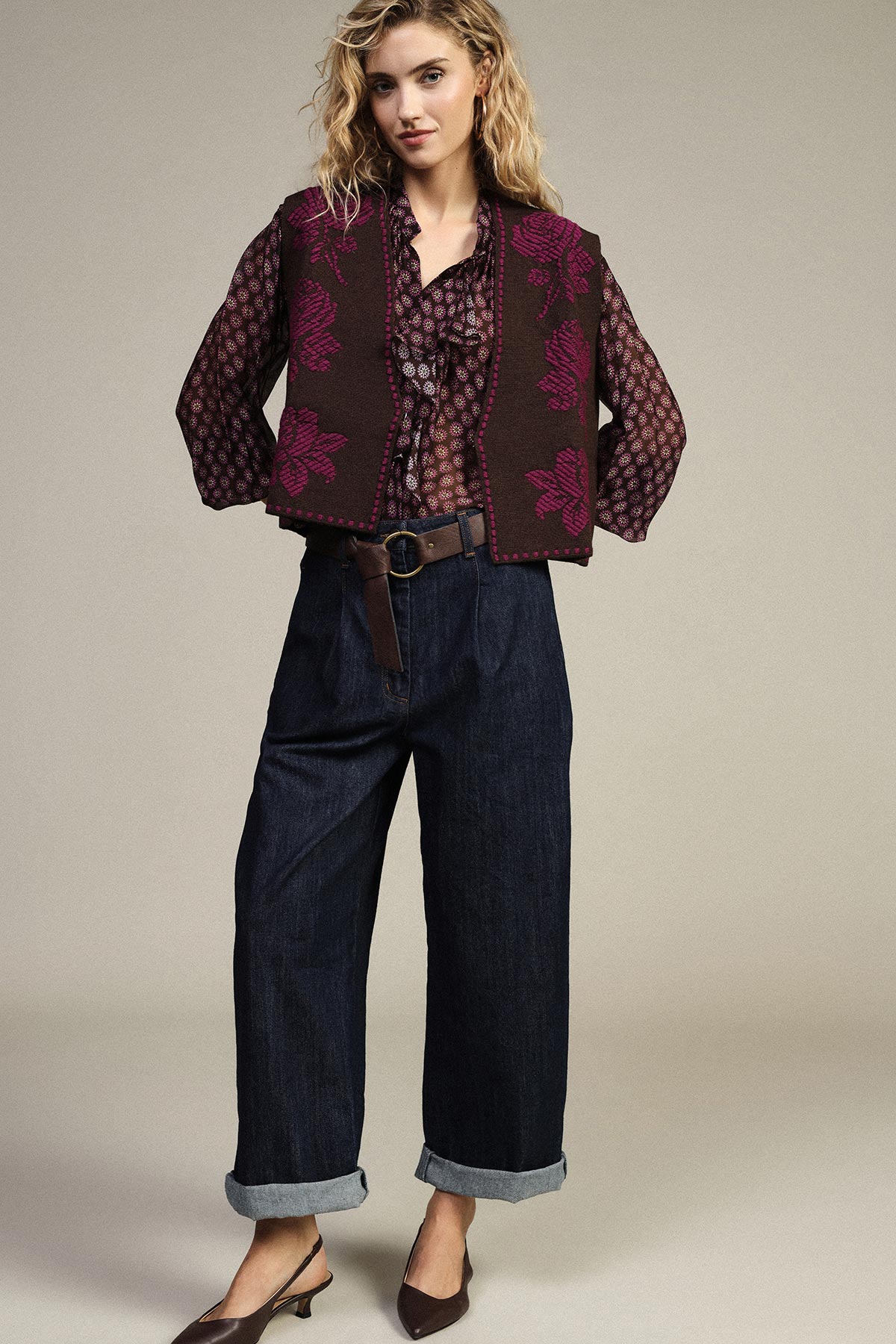 Woman wearing patterned burgundy blouse, knitted floral vest, wide-leg cuffed dark jeans, leather belt and slingback heels