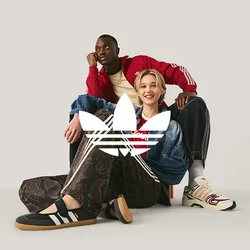 Un uomo con una giacca rossa e una donna con una giacca di jeans e pantaloni Adidas sono seduti sul pavimento davanti a uno sfondo chiaro e uniforme.