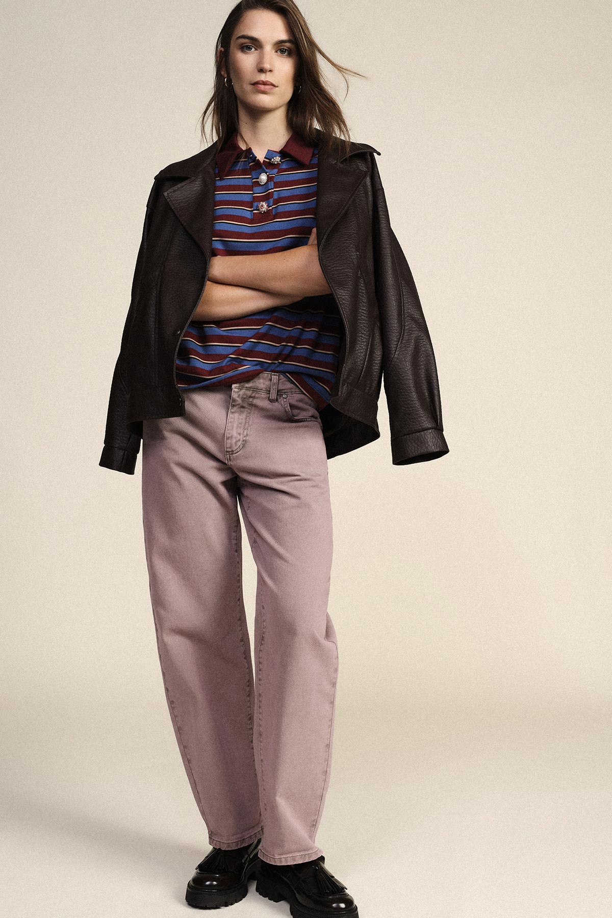 Female model standing with arms crossed wearing brown leather jacket over striped polo, pink wide-leg jeans and black loafers
