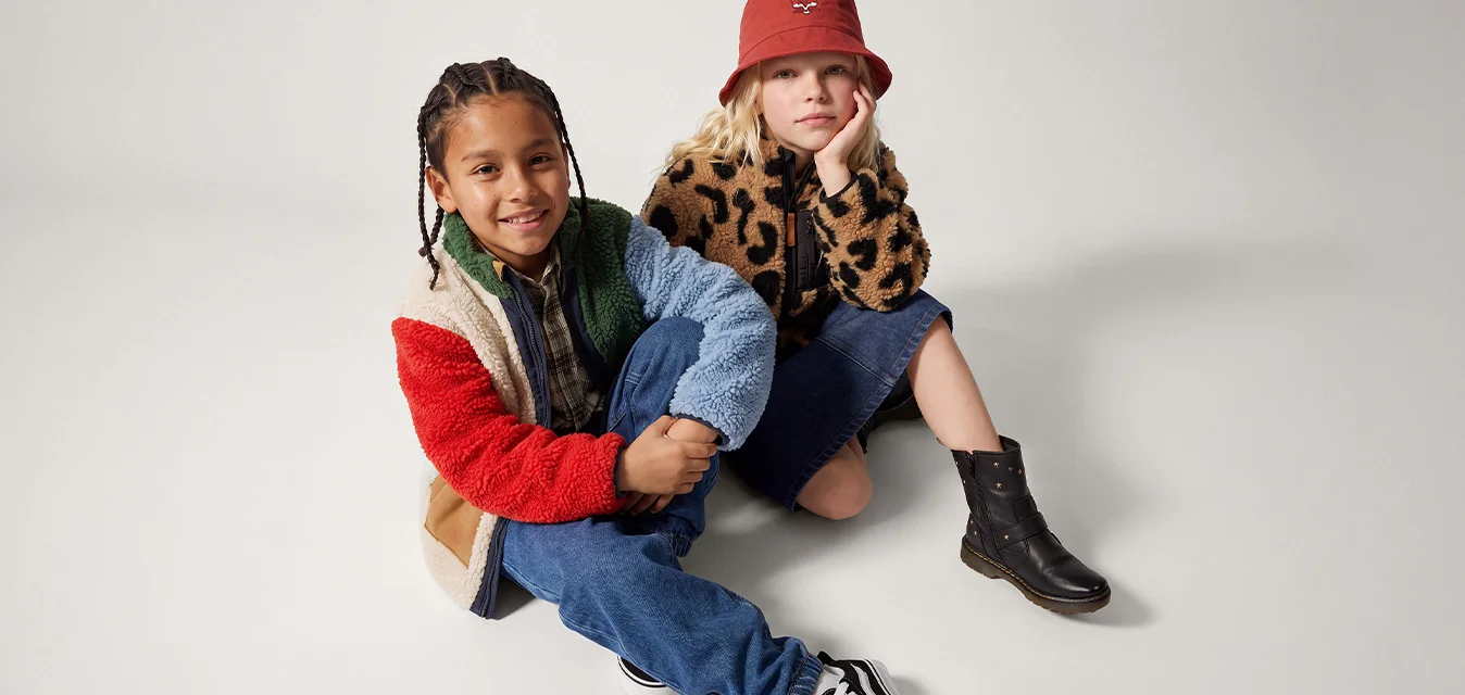 Due bambini seduti sul pavimento, uno con una giacca in pile multicolore e l'altro con una giacca con stampa leopardata e un cappello a secchiello rosso.