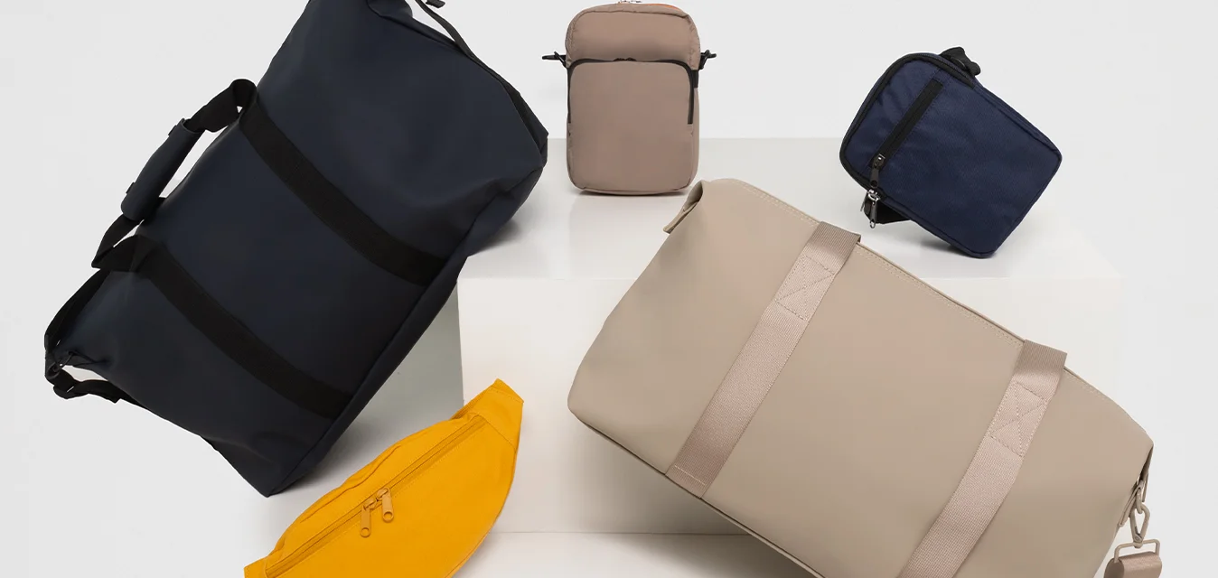 Collezione di borse: borsa da viaggio blu scuro con cinghie, grande borsa da viaggio beige chiaro, marsupio giallo e piccole pochette blu e beige con cerniere.