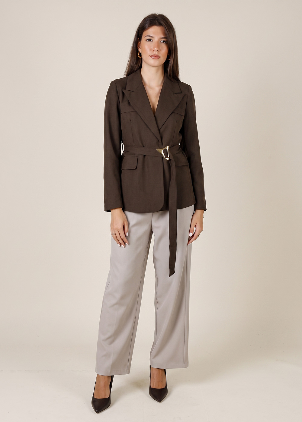 Female model in studio wearing a brown belted blazer with gold buckle and wide-leg beige trousers, black heels, neutral background
