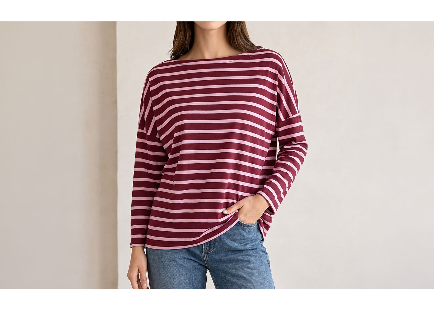 Woman wearing maroon and white striped long-sleeve top and blue jeans, hand in pocket against a neutral background