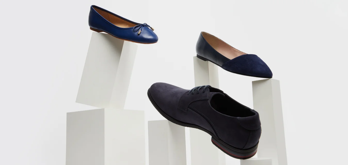 Tre eleganti scarpe scure esposte su piedistalli bianchi inclinati contro uno sfondo chiaro e semplice.