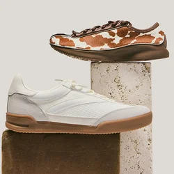 Due sneaker casual esposte su blocchi rettangolari impilati; una bianca con suola marrone chiaro, l'altra marrone con motivo a macchie bianche da mucca.