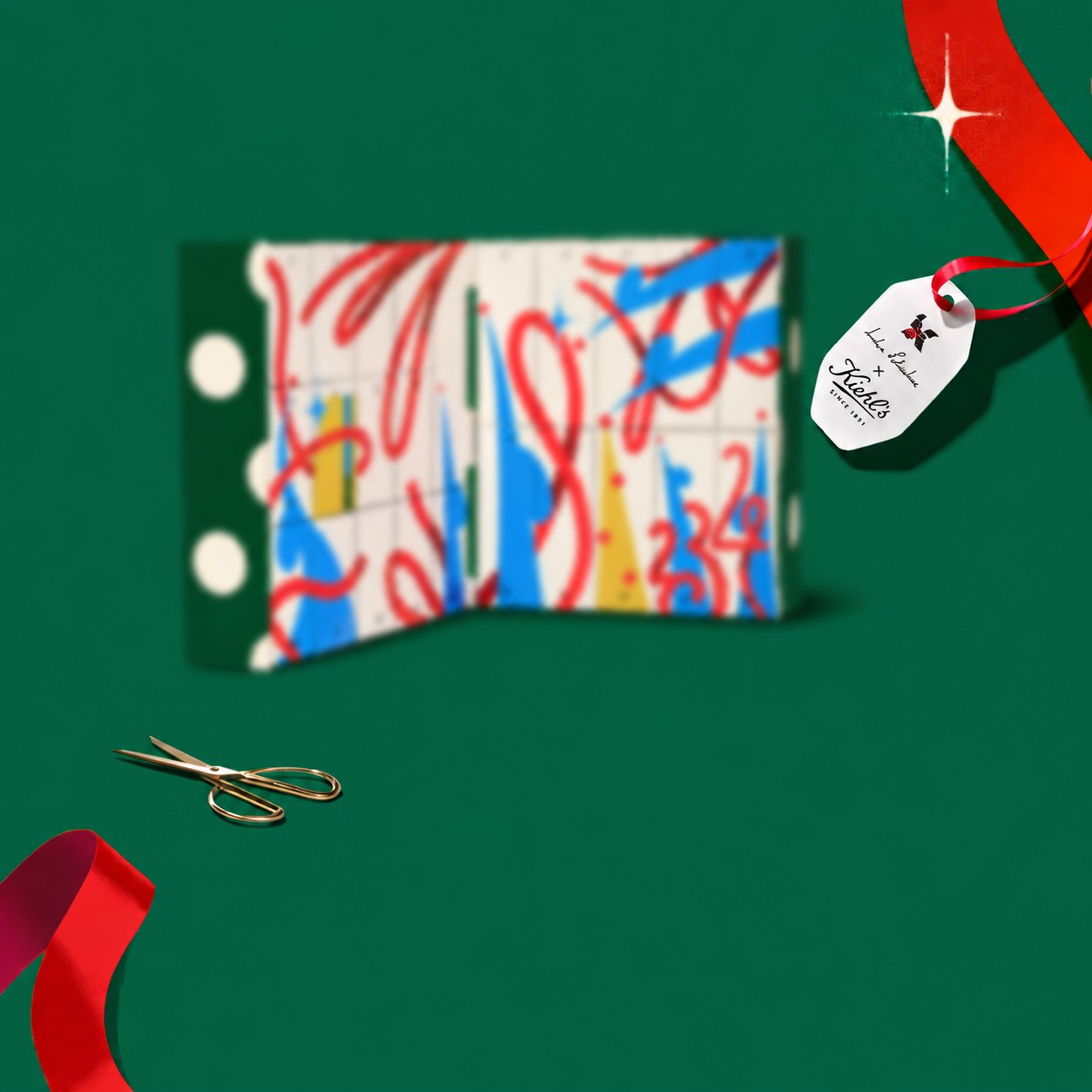 Calendario dell'Avvento Kiehl's 2026 sfocato con etichetta dell'artista, nastri e forbici su sfondo verde
