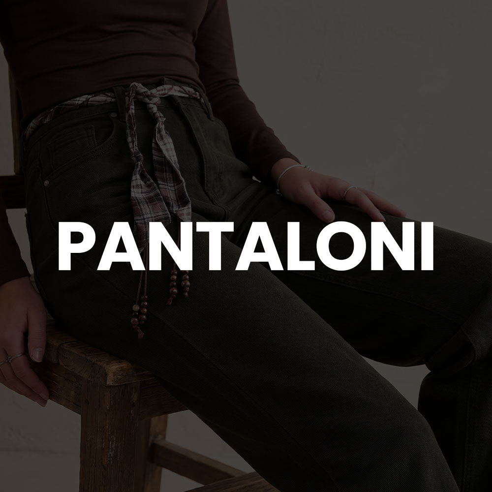 Seated model wearing dark green corduroy trousers with a knotted plaid belt and beaded tassels, hands resting on a wooden stool