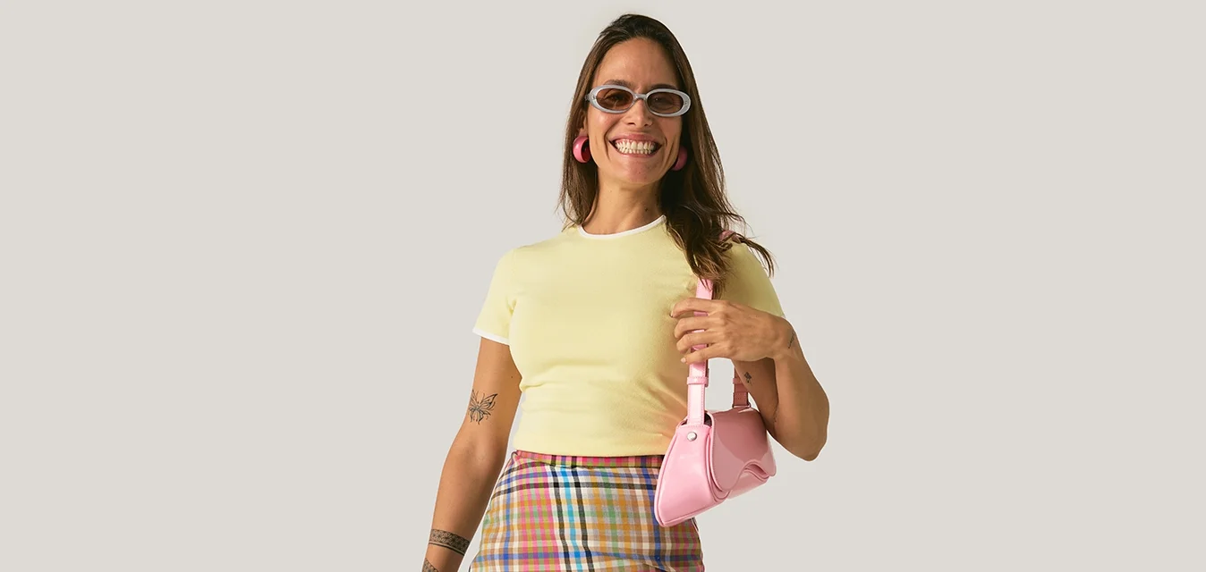 Donna sorridente che indossa un crop top giallo, gonna a quadri, occhiali da sole e tiene una piccola borsa rosa su sfondo chiaro uniforme.