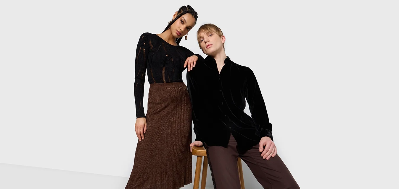 Due modelli posano in uno studio. La donna indossa un top a coste nero e una gonna a coste marrone con stivali fino al ginocchio. L'uomo indossa una camicia di velluto nera e pantaloni marroni.