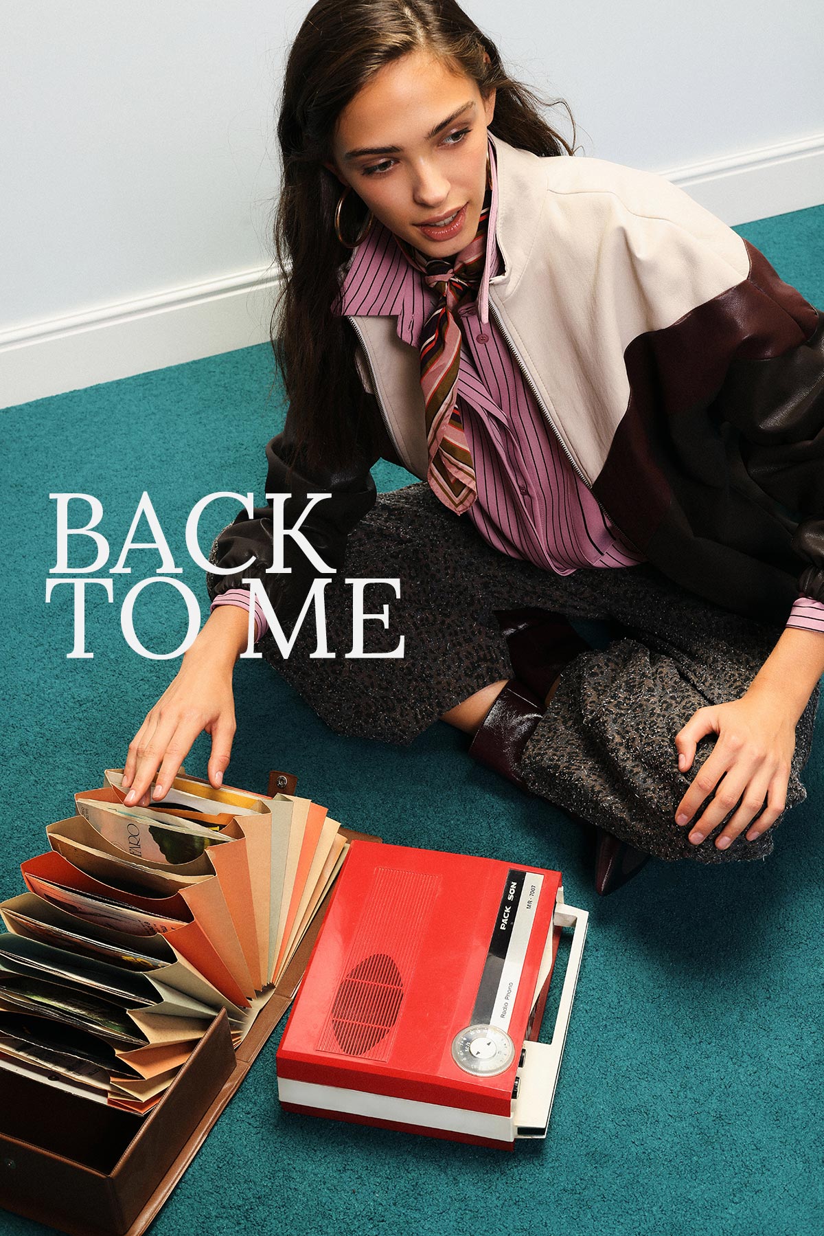 Woman sitting on teal carpet with vintage red cassette recorder and colour-swatch fan, wearing a striped blouse, tie and jacket