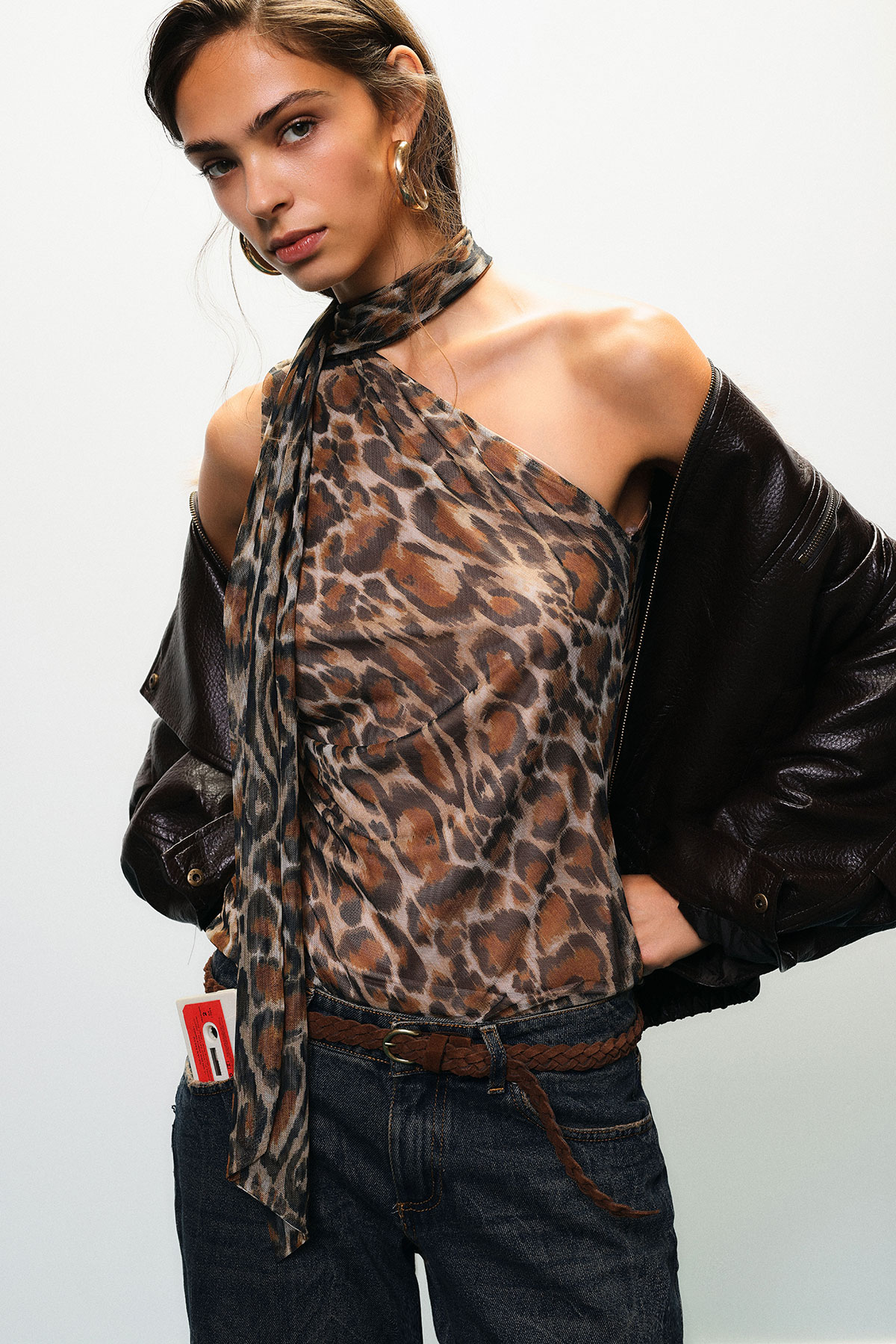 Woman modelling leopard-print halter top with sheer scarf tie, off-shoulder dark leather jacket, hoop earrings and jeans with braided belt
