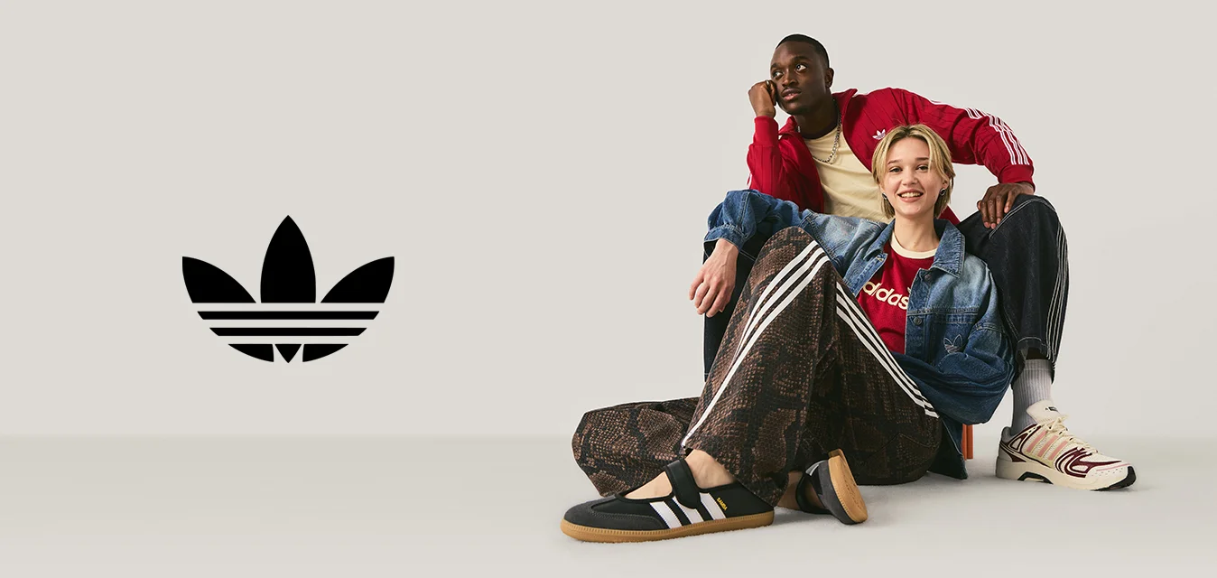 Un uomo con una giacca rossa e una donna con una giacca di jeans e pantaloni Adidas sono seduti sul pavimento davanti a uno sfondo chiaro e uniforme.