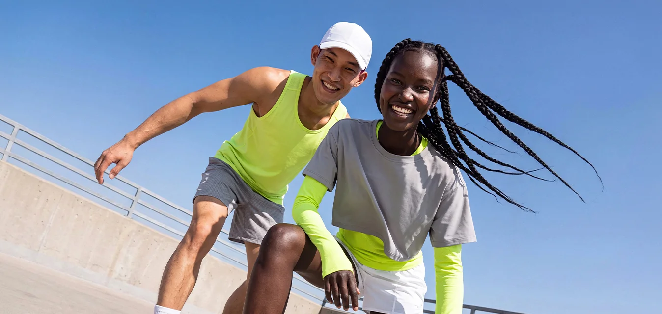 Un uomo e una donna in abbigliamento sportivo si accovacciano e sorridono su un tetto di cemento con un cielo azzurro limpido sopra.